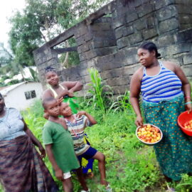 Women and Children in a field with one woman holding bowls of tomatoes.